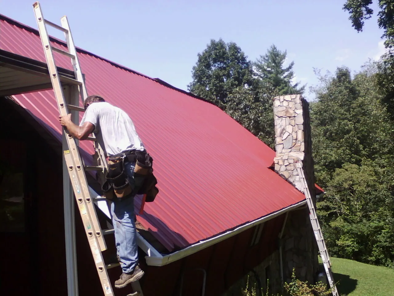 Roofer on a ladder beside a red metal roof and stone chimney — Soffit & Fascia Repair by Roofing Brothers in Bybee